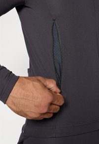 Poche zippée sur une veste d'athlétisme grise foncée, avec une doublure texturée. Main tenant la fermeture éclair, mettant en valeur le tissu lisse et le design.