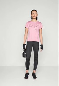 Kvinne med en rosa Puma T-skjorte, svarte leggings, hansker og joggesko, som holder en svart kettlebell i sin høyre hånd, står foran en ensfarget bakgrunn.