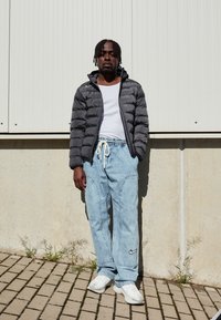 Grå pufferjakke, hvid tanktop, lyseblå jeans med sidelommer og hvide sneakers med grå accenter, poseret mod en ensfarvet baggrund.