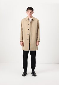 Beige cotton coat with black buttons, long sleeves, and cuff details. Worn over a black top and paired with black tapered pants and shoes.