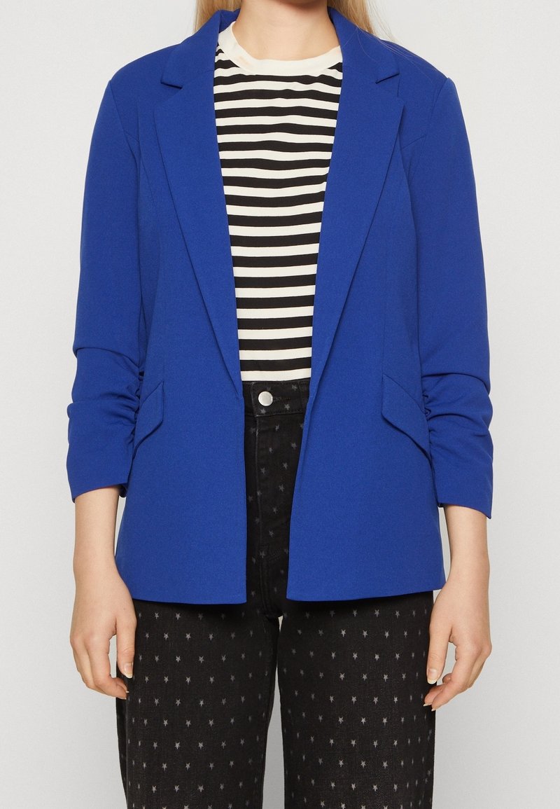 Personne portant un blazer bleu vif sur une chemise rayée noire et blanche avec un pantalon noir présentant de petits motifs en forme d’étoiles.