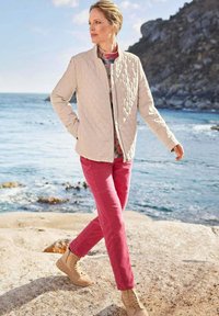 Gequiltede beige jas, gebloemd shirt, roze broek en tan laarzen. Landschapsachtergrond met water en rotsachtige oever. Natuurlijk licht.