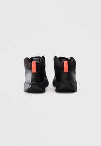 Une paire de baskets noires mi-hautes avec des semelles texturées, des cols de cheville côtelés et des languettes orange vif sur un fond blanc uni.