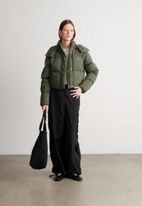 Veste en duvet verte avec un design court et un col oversized, associée à une longue jupe noire et un sac fourre-tout noir. Chaussures plates visibles.