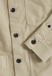 Beige button-up shirt met lange mouwen, voorzien van zwarte knopen, platte voorzakken en stikdetails op de stof.