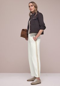 Femme debout de profil portant une veste courte noire, un top beige, un pantalon blanc avec une bande beige, des baskets beiges et tenant un sac marron.