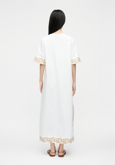Femme aux longs cheveux noirs portant une robe midi blanche avec une bordure beige bouclée aux manches et à l'ourlet, debout pieds nus sur un fond blanc.