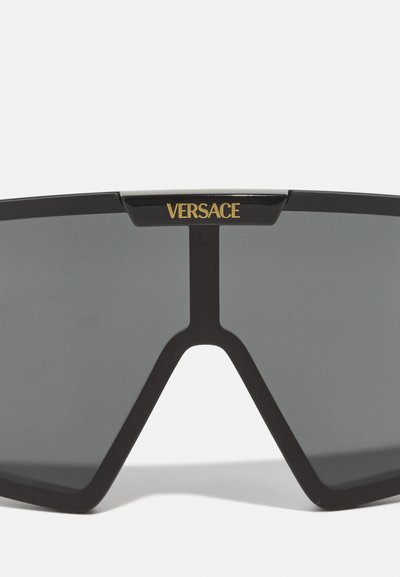 Black sunglasses with a geometric design. Grey lenses. Gold "VERSACE" logo prominently displayed on the top bar. Smooth plastic frame.