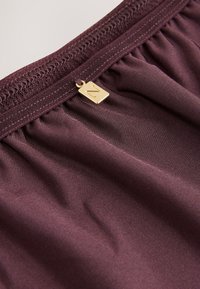 Tissu bordeaux à la texture lisse avec une ceinture élastique, pr�ésentant une petite étiquette dorée portant la lettre « Z » attachée.