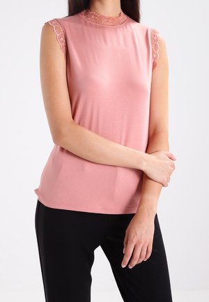 Vrouw draagt een mouwloze roze top met kanten afwerking aan mouwen en kraag, gecombineerd met een zwarte broek, staand met één arm die de andere vasthoudt.