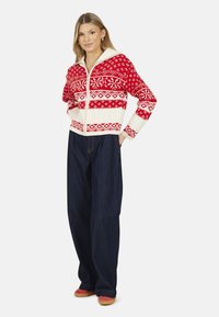 Il cardigan lavorato a maglia presenta un motivo geometrico rosso e bianco, chiusura con zip e un ampio colletto. Abbinato a jeans larghi neri e scarpe rosse.