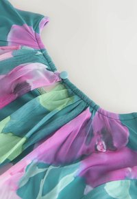 Doorschijnende bloemenblouse met een mix van roze, groen en blauw. Ontwerp met plooien en een turquoise knoop bij de hals voor sluiting.