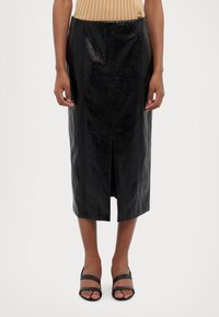 Svart lær midi skjørt med frontspalte, med en blank tekstur og vertikale sømdetaljer, iført enkle svarte sandaler.