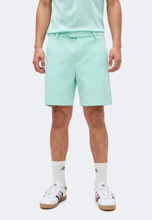 Homme portant un short vert menthe, des chaussettes blanches Adidas et des baskets blanches Adidas avec des bandes noires et rouges, debout devant un fond uni.