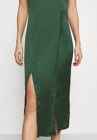 Nainen, joka on pukeutunut hihattomaan tumman vihreään satiiniseen midi-mekkoon, jossa on kaksi etureikää, jotka paljastavat jalat, seisoo valkoista taustaa vasten.