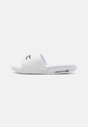 Sandalias deslizantes blancas hechas de material sintético, con suela plana, correa ancha con el logo verde de Lacoste y plantilla texturizada.