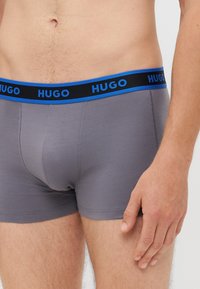 Sive bokserice iz mehkega materiala, z modrim in črnim elastičnim pasom s logotipom "HUGO", prilegajoča zasnova in gladka tekstura.