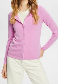 Vaaleanpunainen pehmeästä kankaasta valmistettu neulepusero, jossa on napitettava etuosa. Mallissa on V-pääntie, pitkät hihat ja istuva leikkaus. Yhdistetty vaaleiden housujen kanssa.