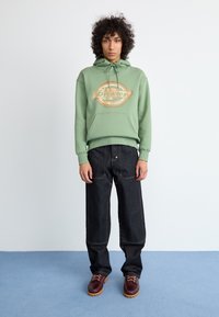 Sweatshirt verde claro com um logotipo central, jeans largosos pretos e sapatos borgonha com solas castanhas. Textura suave e ajuste descontraído.