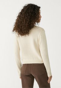 Kremowy sweter dzianinowy z długimi rękawami, z prążkowanymi mankietami i dołem. W zestawieniu z brązowymi spodniami. Teksturowana powierzchnia i luźny krój.