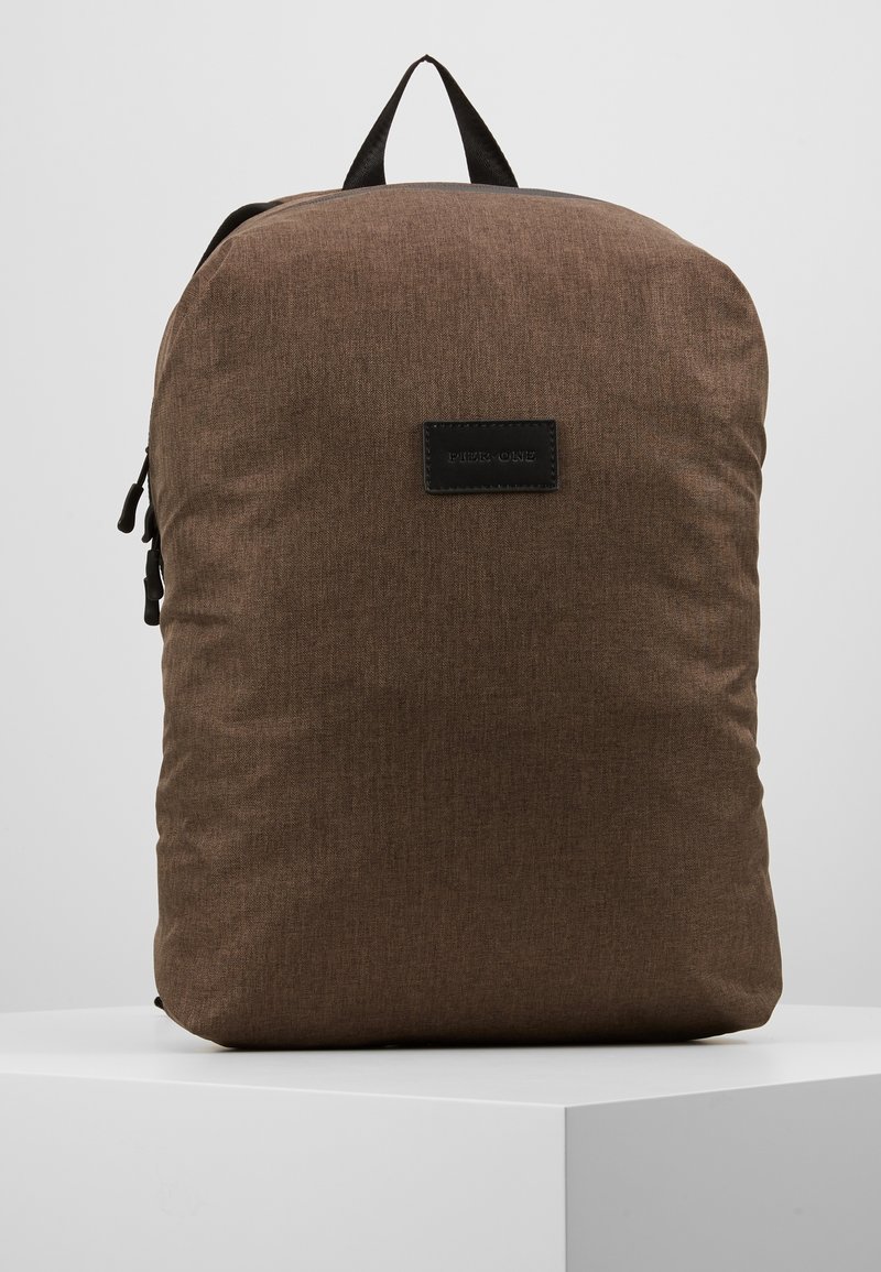 Sac à dos en tissu brun avec une texture lisse, doté d'une étiquette en cuir noir et d'une poignée de transport en haut. Design simple et minimal.