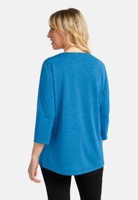 Blonde vrouw in een felblauw langslavend top en zwarte broek, staand, met haar rug naar de camera, tegen een effen witte achtergrond.