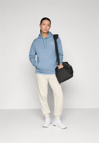 Hellblaue Kapuzenjacke mit einer Fronttasche und Kordelzug; kombiniert mit cremefarbenen Jogginghosen und weißen Turnschuhen; hält eine schwarze Sporttasche.