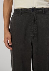 Pantalones de pana negros con canalones verticales, un bolsillo frontal y un cierre de botón. La tela parece suave con una superficie texturada.