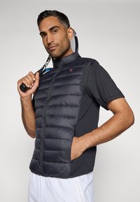 Δεμένο γιλέκο χωρίς μανίκια σε σκούρο γκρι με μπροστινό φερμουάρ, σε συνδυασμό με μαύρο μπλουζάκι κοντομάνικο. Μια ρακέτα τένις κρατείται σε μία χέρι.