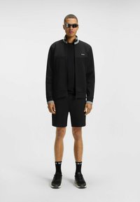 Giacca nera con zip e collo a strisce, abbinata a pantaloni corti neri. Gli accessori includono occhiali da sole riflettenti e scarpe da ginnastica nere con calze logate.