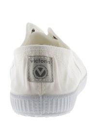 Zapatilla blanca tipo slip-on con suela de goma texturizada y etiqueta de logo gris en el talón. Presenta costuras simples y acentos elásticos en los laterales.