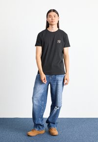 Tricou negru cu mânecă scurtă, având un imprimeu grafic, asortat cu blugi deschiși la culoare, uzati, și teniși maro. Design simplu și casual.