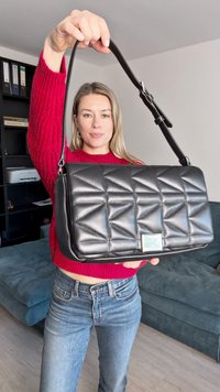 Γυναίκα με κόκκινο πουλόβερ κρατά μια μαύρη καπιτονέ δερμάτινη τσάντα με λουρί ώμου σε ένα σαλόνι.