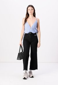 Blouse bleue à motifs avec un décolleté en V profond, nouée sur les côtés ; jeans noirs taille haute ; sac à main noir ; baskets de couleur claire.