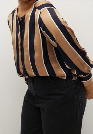 Personne portant un chemisier à manches longues avec des rayures verticales beige, noires et blanches, associé à un pantalon noir taille haute.