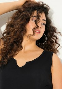 Zwarte geribbelde tanktop met een V-hals, voorzien van brede schouderbanden en een gestructureerde stof. Het model heeft krullend haar en oorbellen met ringen.