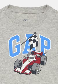 Γκρι βαμβακερό μπλουζάκι με γραφικό ενός κόκκινου αγωνιστικού αυτοκινήτου και μια σημαία με καρό σχέδιο, με το "GAP" σε έντονα μπλε γράμματα πάνω από αυτό.