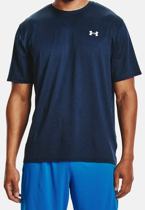 Mężczyzna ubrany w granatową koszulkę Under Armour z krótkim rękawem i jaskrawo niebieskie krótkie spodenki sportowe, stoi z rękami swobodnie opartymi po bokach.