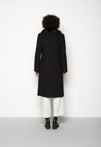 Largo abrigo negro con cuello de pelo sintético, diseño entallado, puños abotonados y corte recto, complementado con pantalones de pierna ancha blancos.