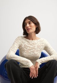 Långärmad topp gjord av texturerad, krämfärgad tyg med utskurna mönster. Parat med mörka jeans, mot en blå bakgrund.