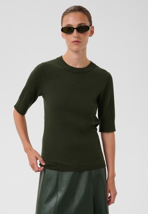 Femme aux cheveux plaqués en arrière portant des lunettes de soleil foncées, un pull côtelé à manches courtes vert foncé et une jupe en cuir vert foncé sur fond gris.