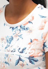 Zbliżenie na szyję i górną część torsu osoby w białej koszuli w kwiaty z niebieskimi i brzoskwiniowymi kwiatami oraz częściowym białym tekstem pisanym kursywą.