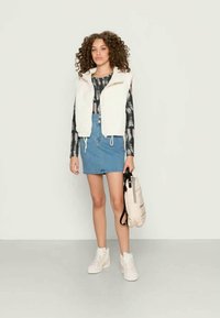 Jovem mulher com cabelo encaracolado, vestindo um colete sem mangas branco, uma camisa de manga longa com padrão tie-dye, uma saia de jeans azul e ténis brancos, segurando uma mochila bege.