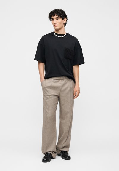 Άνδρας με σγουρά μαλλιά, φορεί μαύρο φαρδύ μπλουζάκι με υφή στην τσέπη, μπεζ παντελόνι με φαρδιά ποδιά, μαύρα παπούτσια και λευκό κολιέ με χάντρες, χέρια στις τσέπες.