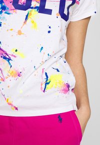 Bílý bavlněný tričko s designem barevného kapání barvy a nápisem "GOLD" tučně napsaným námořnickou modří; spárovány s jasně růžovými šortkami s logem.