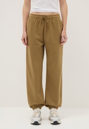 Persona vistiendo pantalones joggers holgados marrones con puños y cintura elásticos, combinados con zapatillas blancas y una camiseta corta blanca.