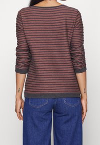 Persona che indossa una maglietta a righe marroni e nere a maniche lunghe con i polsini arrotolati e jeans in denim blu, vista da dietro.