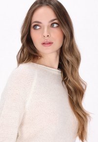 Jasno beżowy sweter z dzianiny o fakturowanym wykończeniu, okrągłym dekolcie i rękawach raglanowych, noszony przez modelkę z długimi, falistymi włosami.