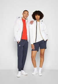 Două modele stau unul lângă altul. Unul poartă o jachetă impermeabilă albă cu accente negre și galbene, un tricou roșu și pantaloni scurți navy. Celălalt poartă o jachetă similară albă peste un tricou alb și pantaloni scurți navy. Amândoi poartă adidași albi.