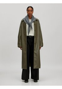 Olijf groene leren jas, knie-lengte, met een knoopsluiting en brede mouwen, gecombineerd met een grijze sjaal en zwarte wijde broek.